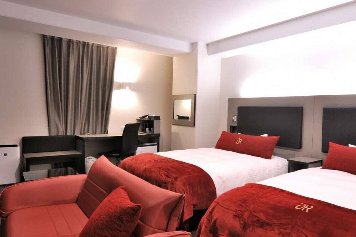 A hotel room with two beds, a desk, a chair, a lamp, and a window with curtains.