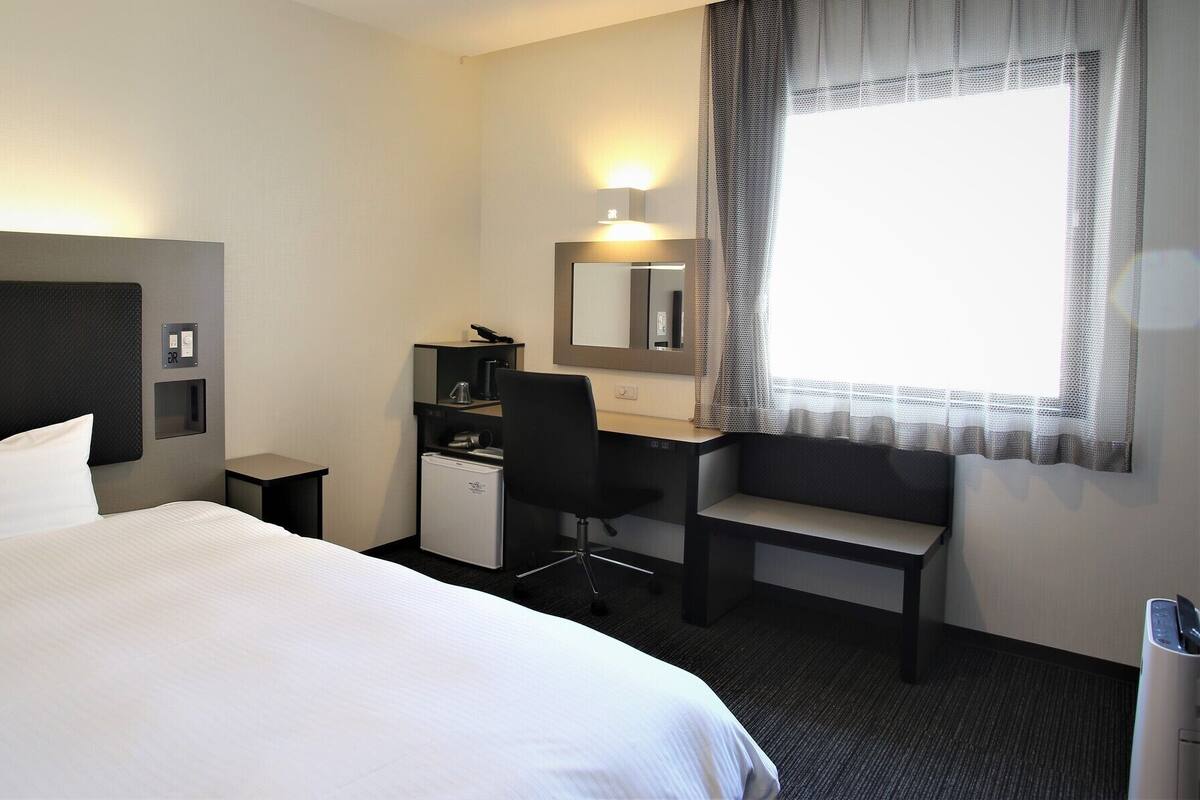 A hotel room with a bed, a desk, a chair, a small refrigerator, a mirror, and a window with sheer curtains.