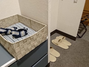 A corner of a hotel room with a woven basket containing a striped bag and a pair of beige slippers on the floor. - Hotel Oaks Early-Bird Osaka Morinomiya (Osaka)