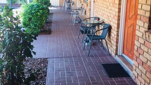 Un patio techado al aire libre con piso de ladrillo, mesas y sillas, y una pared de ladrillo con ventanas y una puerta.