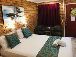 A hotel room with a brick wall, a bed with a patterned bedspread, two chairs, a small table, and a painting.
