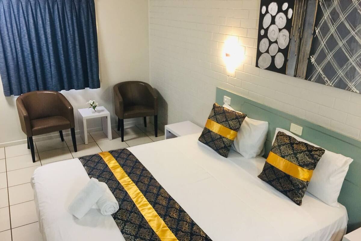 A hotel room with a bed, two chairs, a small table, and a wall-mounted light.
