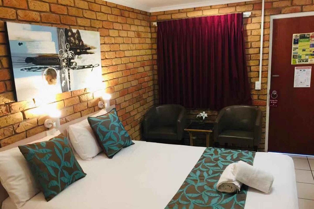 Quarto de hotel com parede de tijolos, cama com colcha estampada, duas cadeiras, uma mesa pequena e um quadro.
