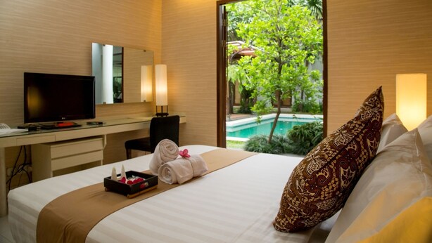 A hotel room with a bed, a desk with a computer, a TV, and a view of a swimming pool and greenery. - Chic Quarter Residence (Jakarta)