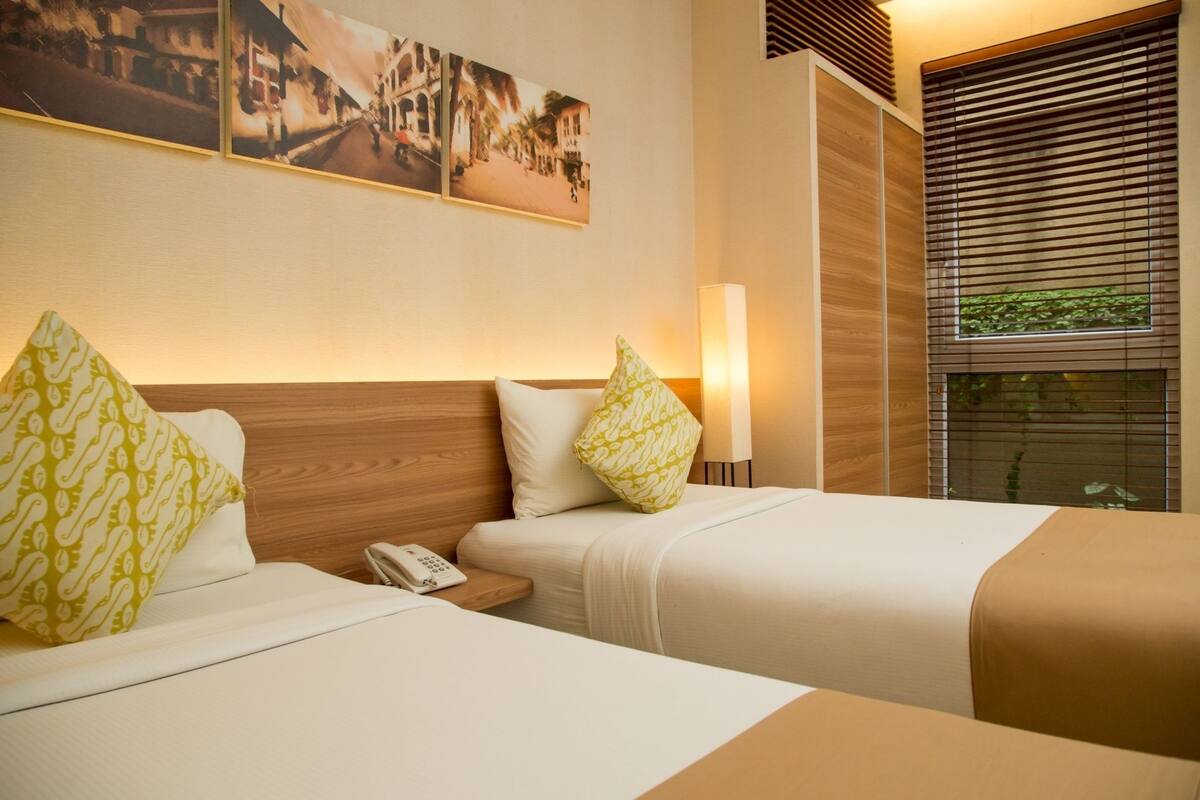 Een hotelkamer met twee bedden, houten hoofdeinden, een nachtkastje met een telefoon en een raam met luxaflex.