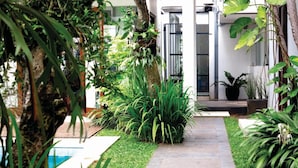 A pathway leading to a modern building with a glass door, surrounded by lush greenery and a small pool. - Chic Quarter Residence (Jakarta)