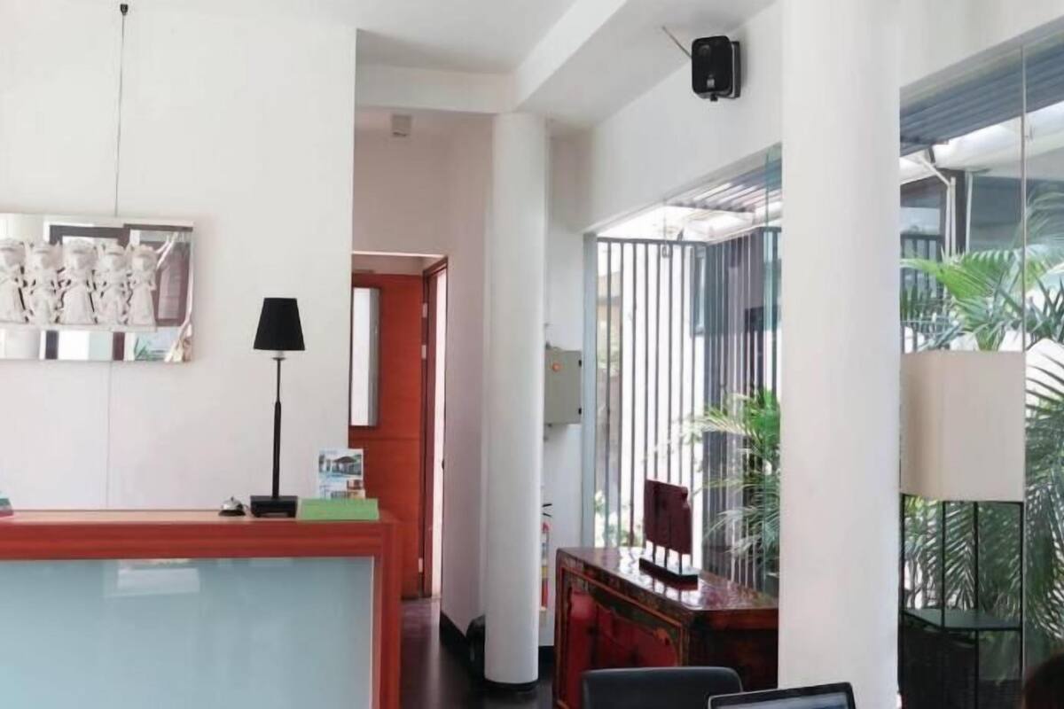 Een hotel receptie met een balie, stoelen, een lamp en een groot raam met uitzicht op palmbomen.