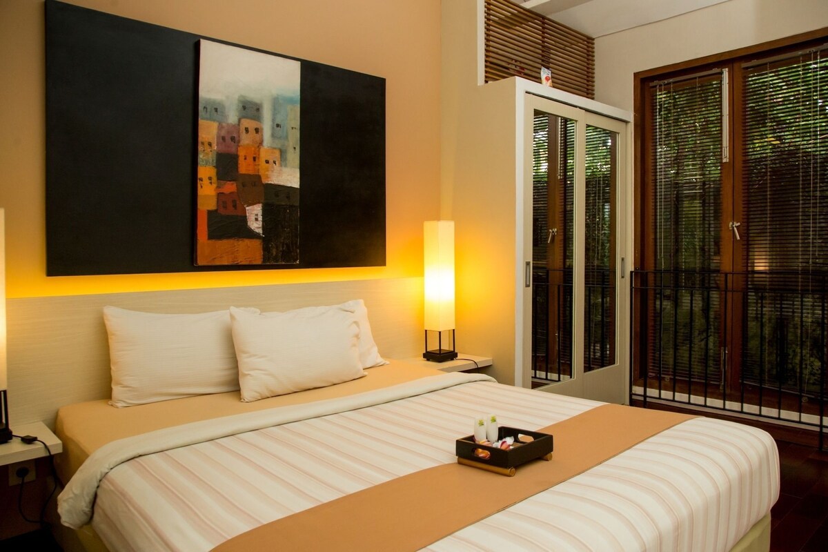 A hotel room with a bed, a nightstand, a painting, a bedside lamp, a tray with toiletries, and a sliding door with a view.