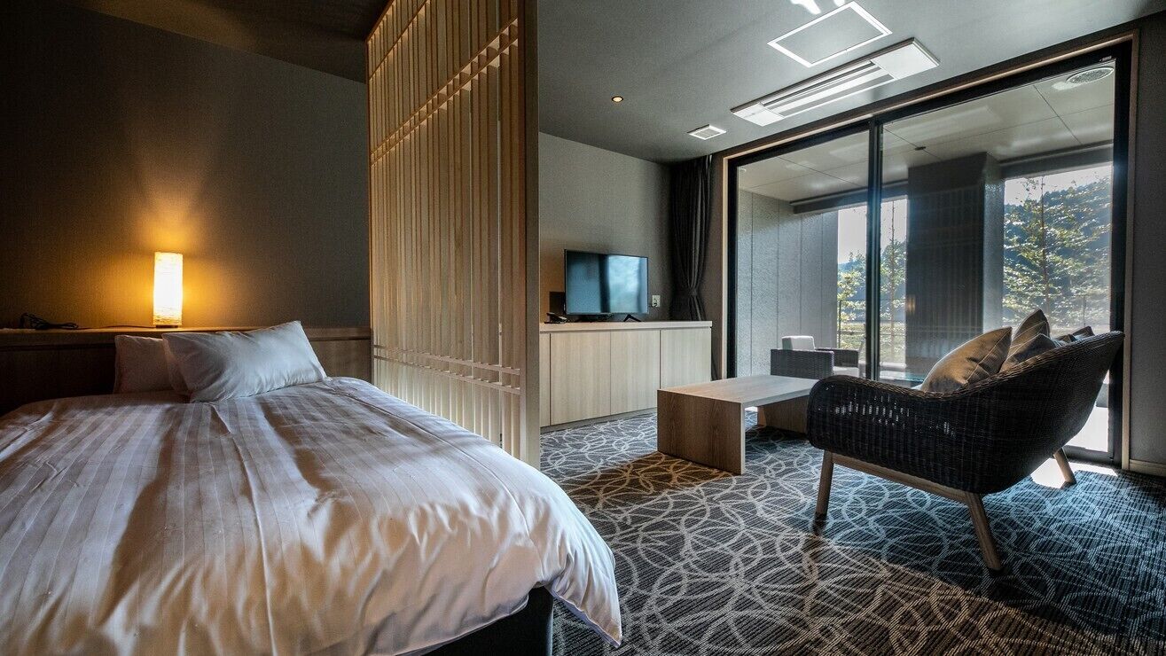 A modern hotel room with a bed, a TV, a seating area, and a large window.