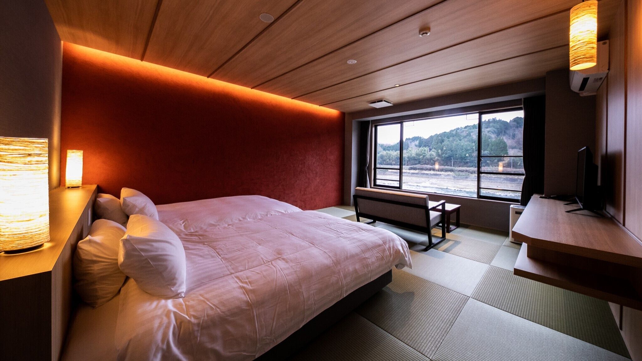 A modern hotel room with a bed, bedside table, lamp, and a large window offering a view of the outdoors.