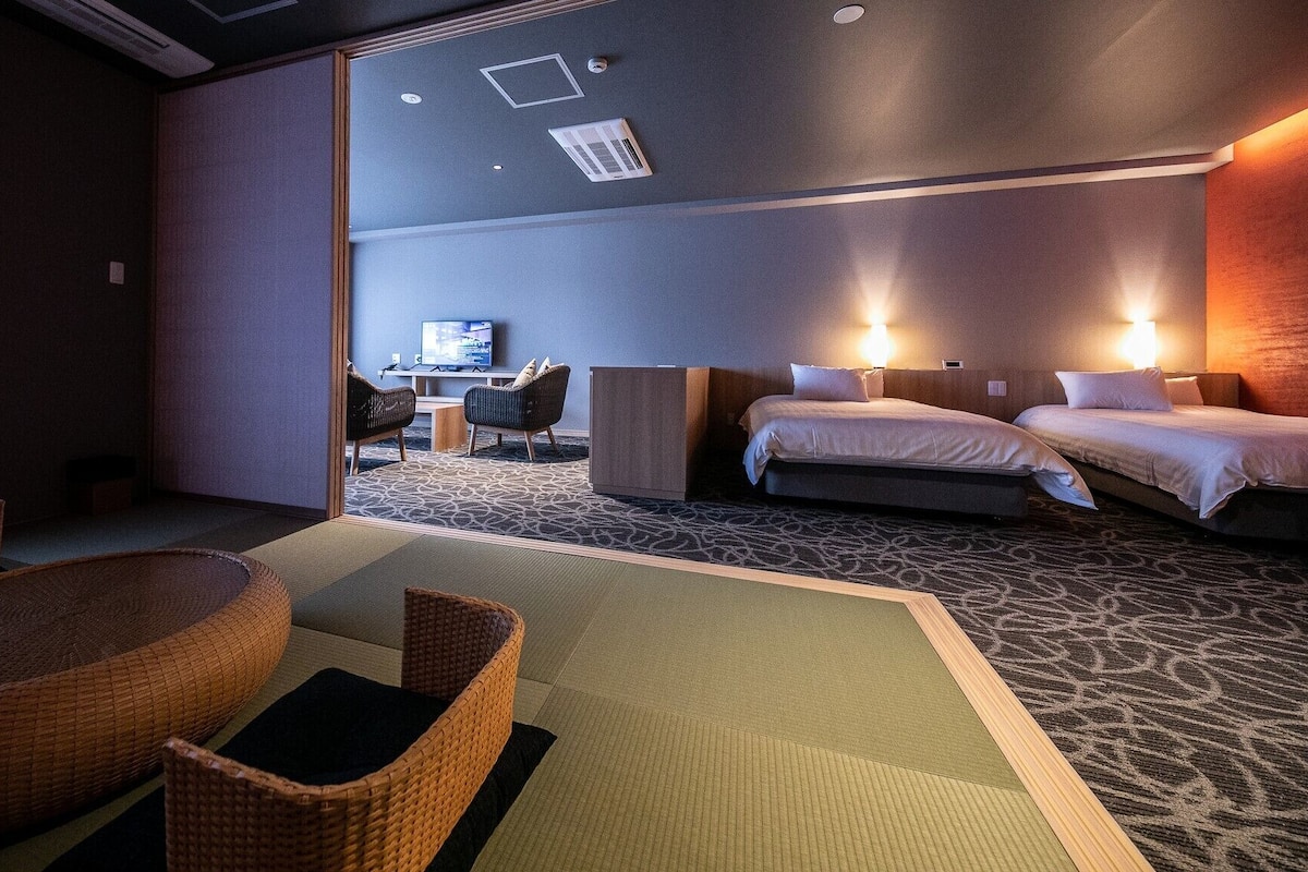 Habitación de hotel con dos camas, televisión, escritorio y zona de estar con una silla y una mesita.