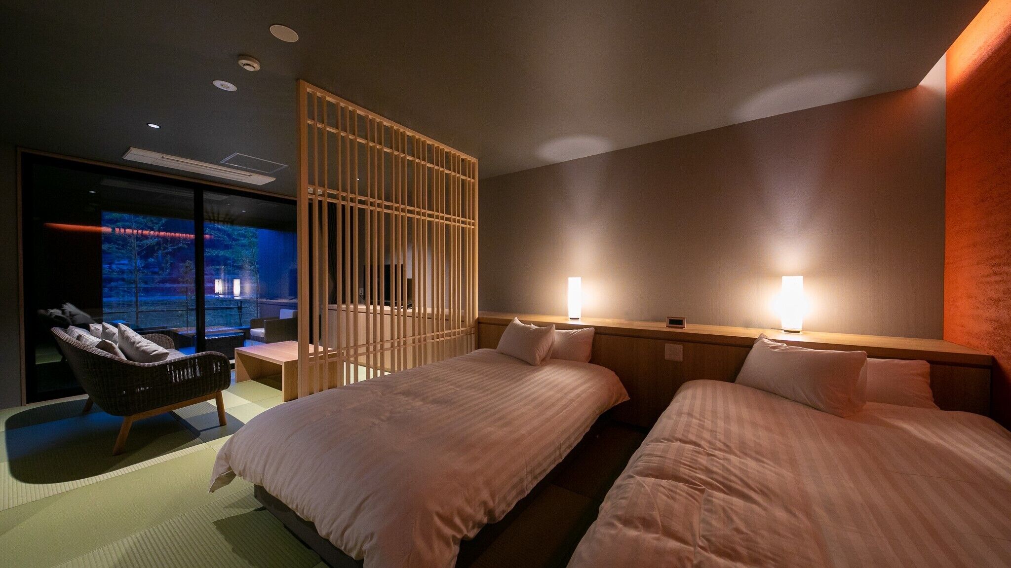 Habitación de hotel con dos camas, un área de descanso con una silla, un ventanal grande y un televisor montado en la pared.