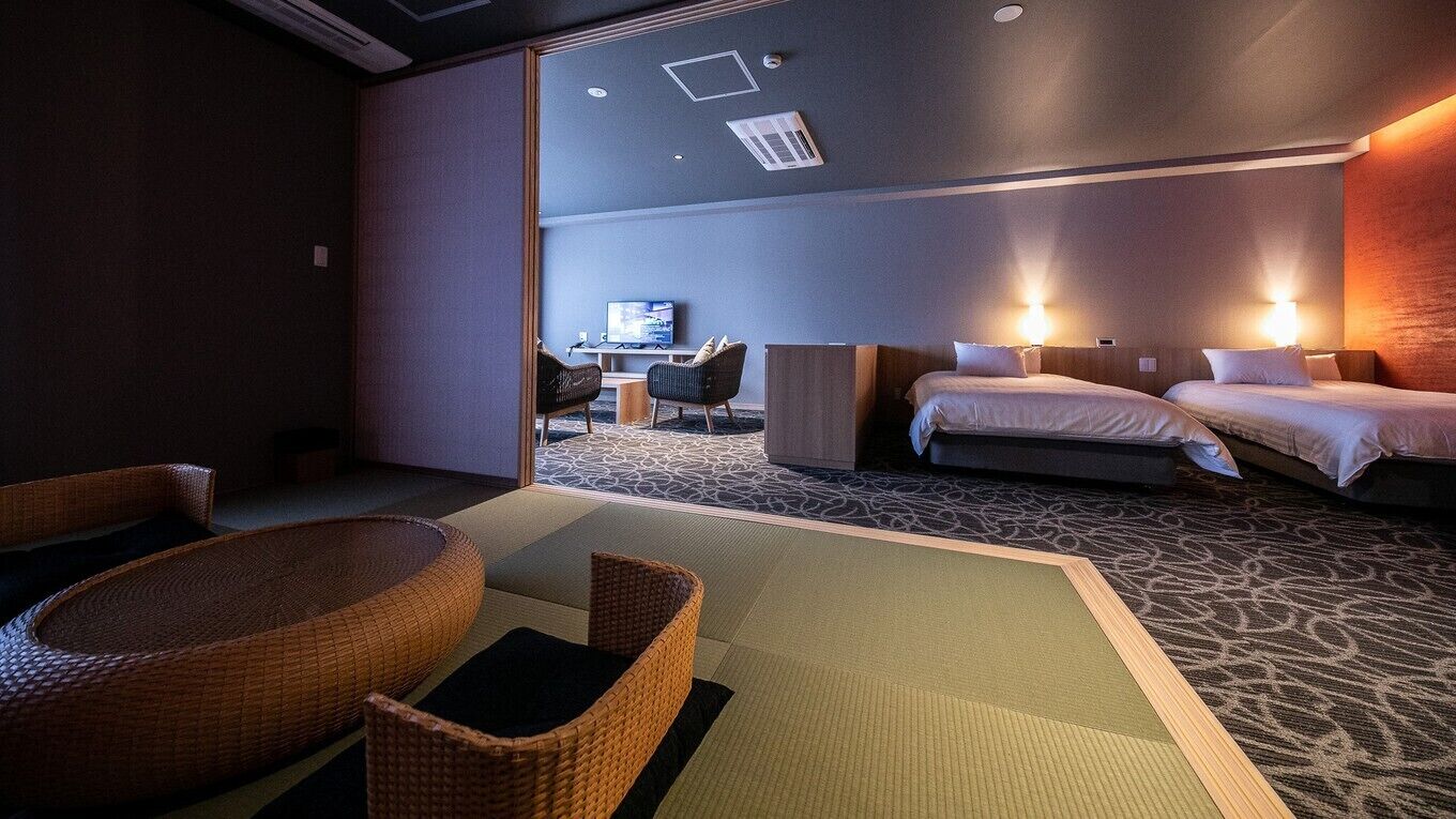 Una habitación de hotel con una cama, dos mesitas de noche, un televisor y una zona de estar con una silla y una mesa baja.
