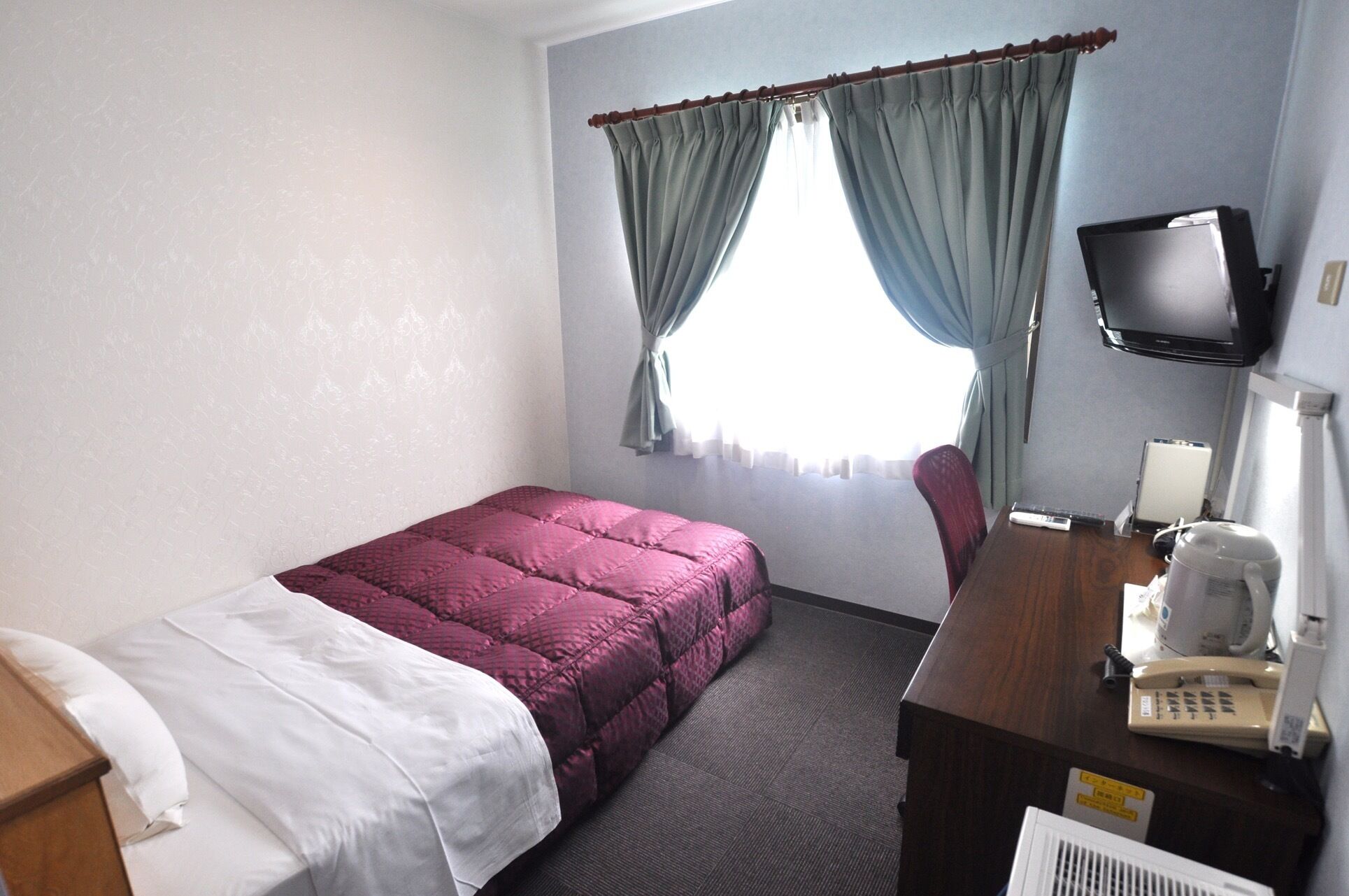 Een hotelkamer met een bed, een bureau met telefoon, een televisie en een raam met gordijnen.