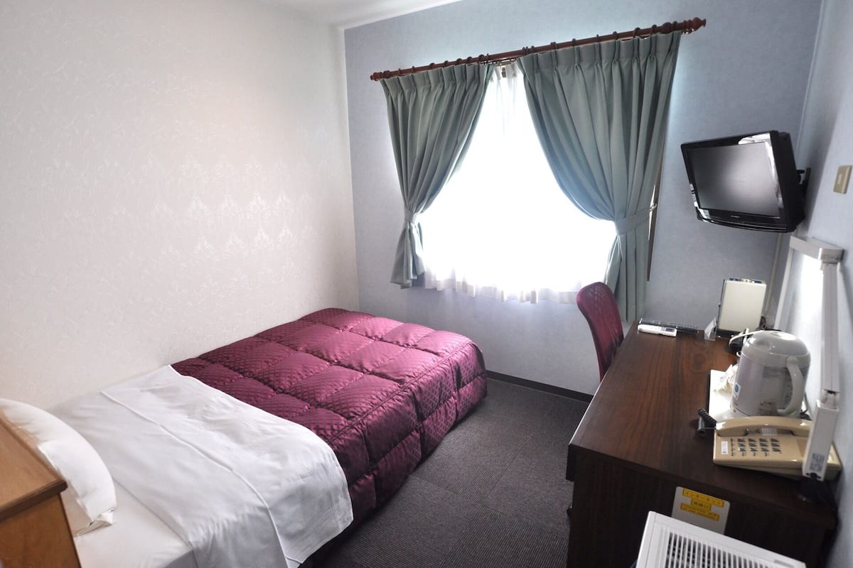 Een hotelkamer met een bed, een bureau met telefoon, een televisie en een raam met gordijnen.