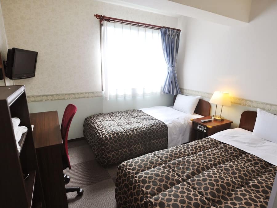 Habitación de hotel con dos camas, un escritorio con silla, televisor y ventana con cortinas.