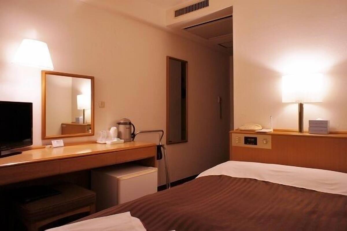 Una habitación de hotel con una cama, un escritorio con lámpara, un televisor y un espejo.