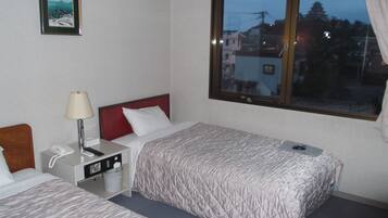 Habitación de hotel con dos camas, una mesita de noche con lámpara, teléfono y ventana con vistas a edificios.