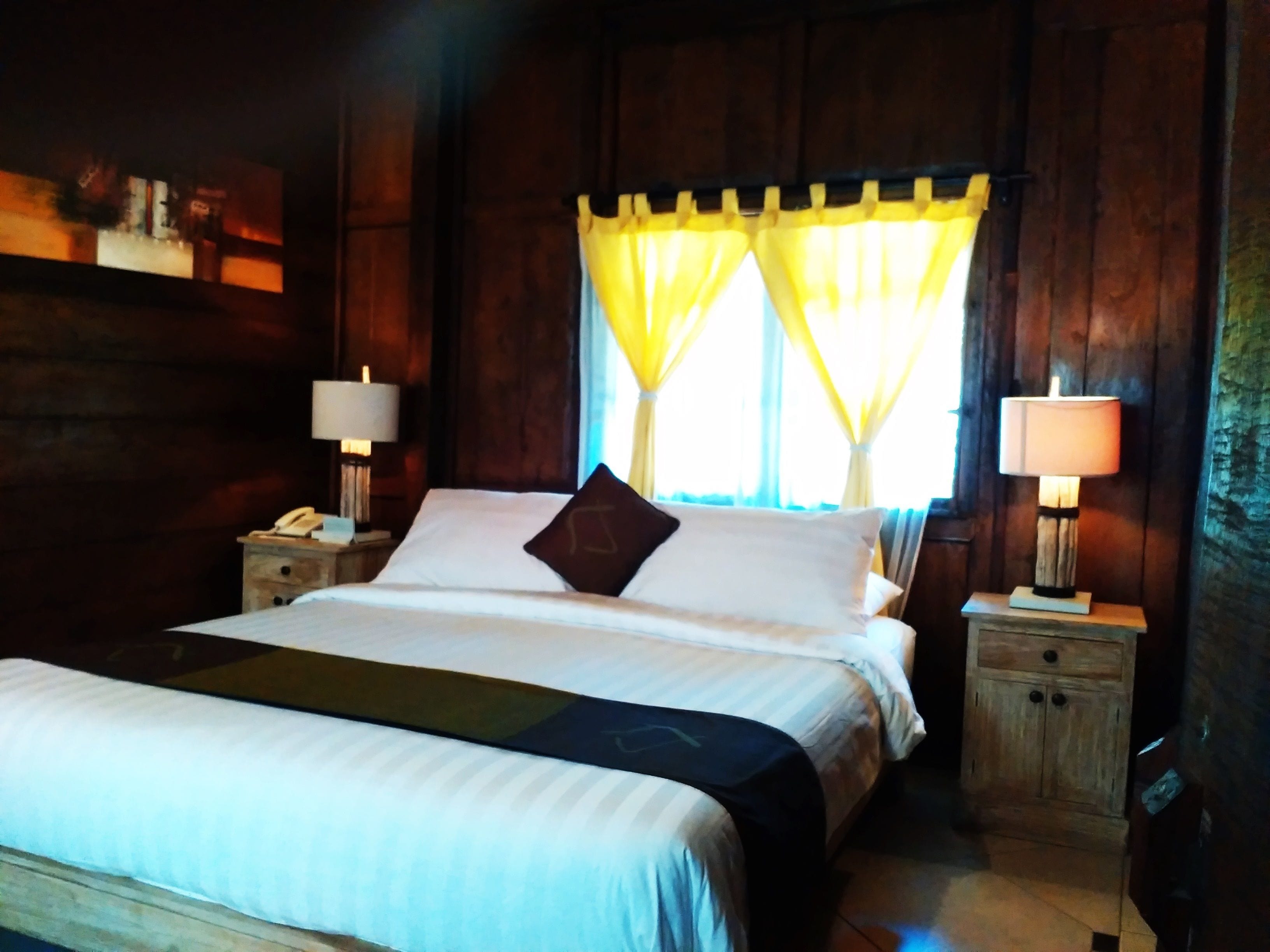A wooden-paneled room with a bed, two bedside tables, and a window with yellow curtains.