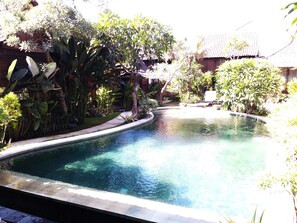A swimming pool with a curved edge, surrounded by lush greenery and a traditional building with a tiled roof. - Kampung Cenik (Legian)