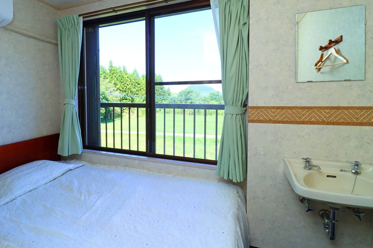 Een kamer met een bed, een raam met gordijnen, uitzicht op groen, een wastafel aan de muur en een handdoekhouder.