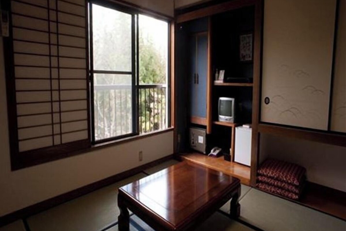 Une pièce de style japonais traditionnel, avec un sol recouvert de tatamis, une petite table en bois, une porte coulissante et un téléviseur posé sur une étagère.