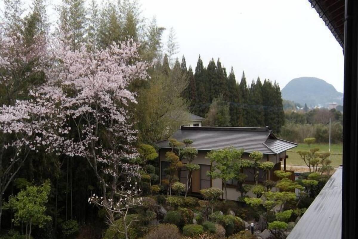 Een traditioneel Japans gebouw met een schuin dak, omgeven door een verzorgde tuin met verschillende struiken en een kersenboom.