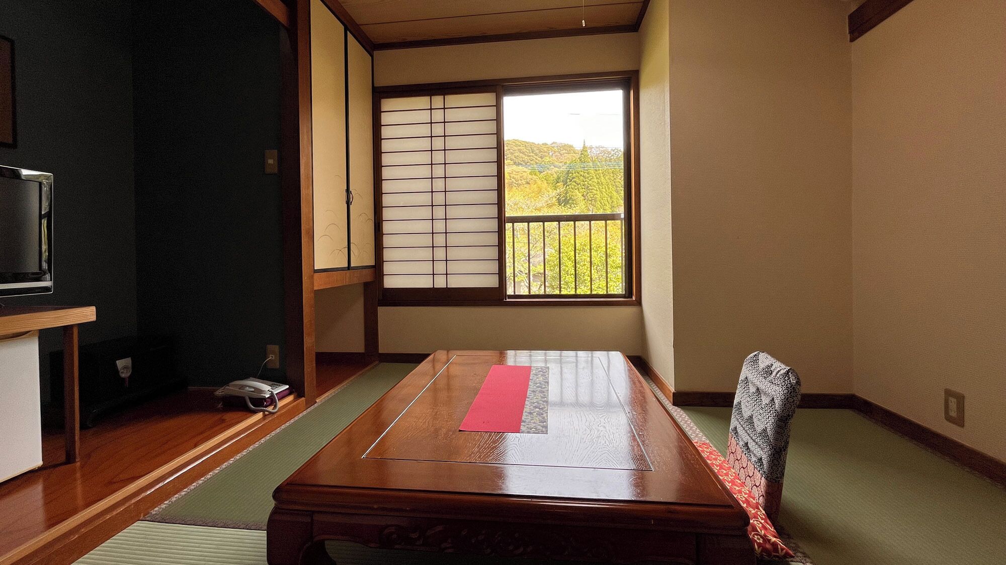 A traditional Japanese room with tatami flooring, a low wooden table, a sliding shoji screen, and a view of greenery outside.