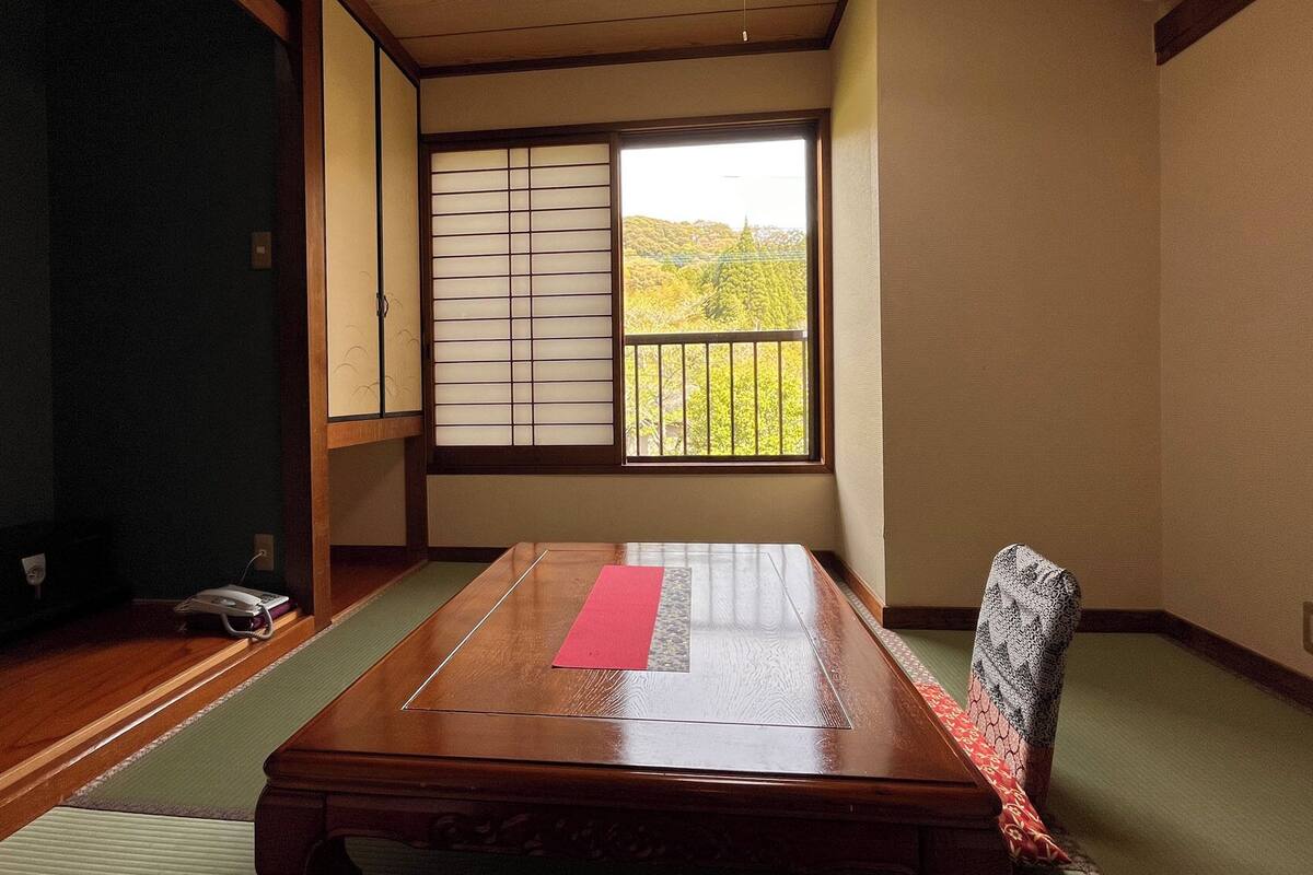 Een traditionele Japanse kamer met tatami vloerbedekking, een lage houten tafel, een verschuifbare shoji-scherm en uitzicht op groen buiten.