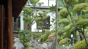A traditional Japanese garden with a stone path, various shrubs, and a building with large windows in the background. - Ryokan Satsumanosato (Satsumasendai)