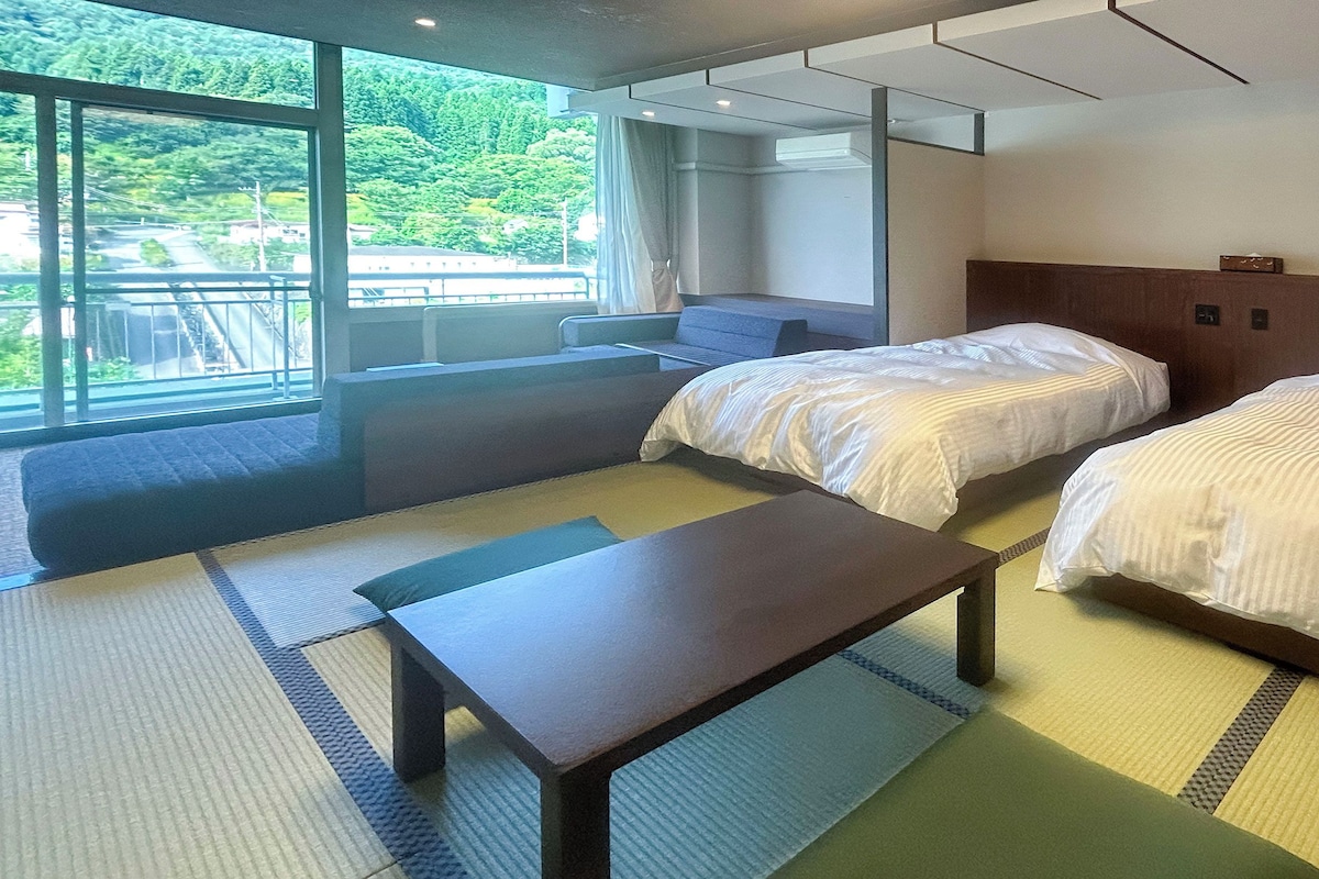 A hotel room with two beds, a small table, and a view of a green landscape through large windows.