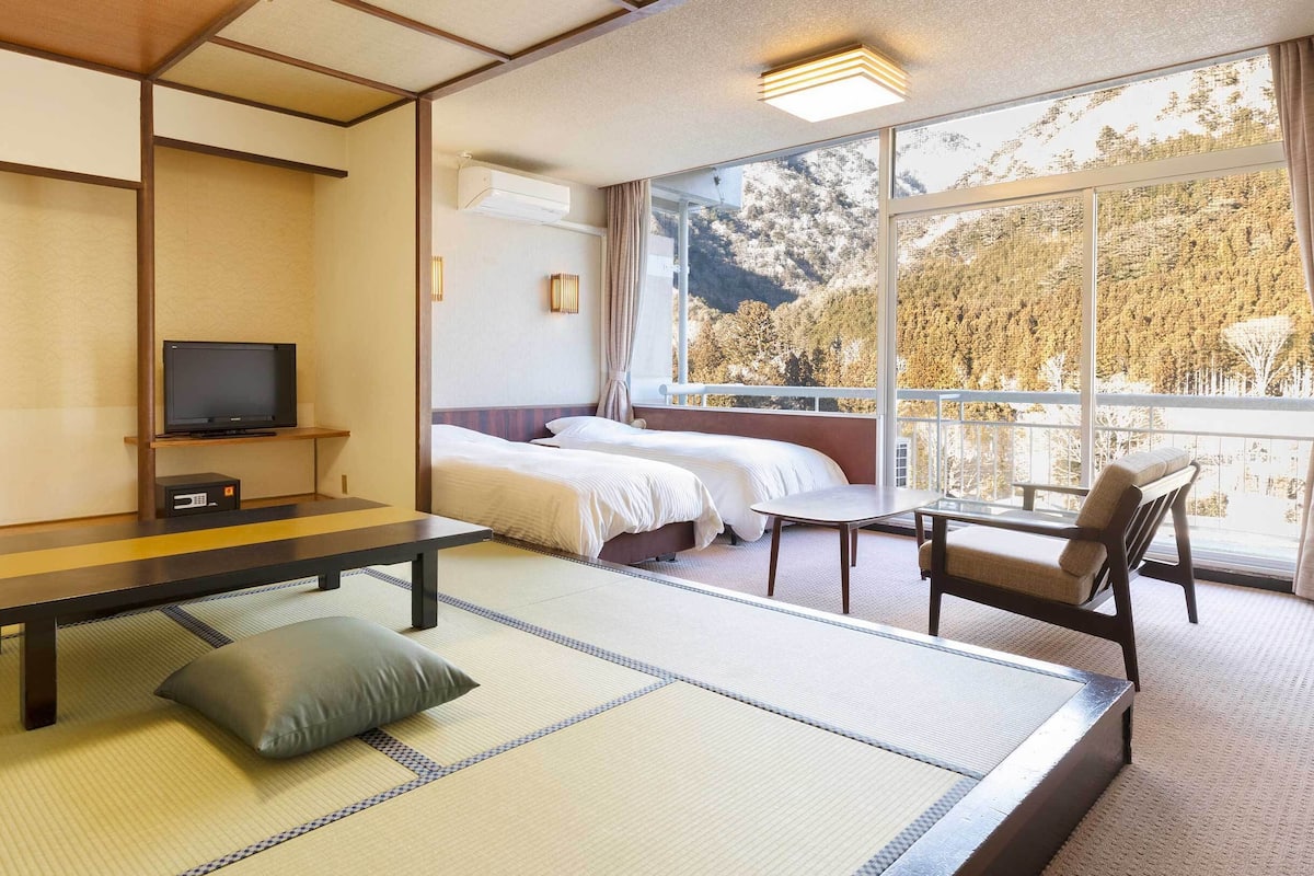 Una habitación con suelo de tatami, televisión, dos camas, sofá y balcón con vistas a árboles.