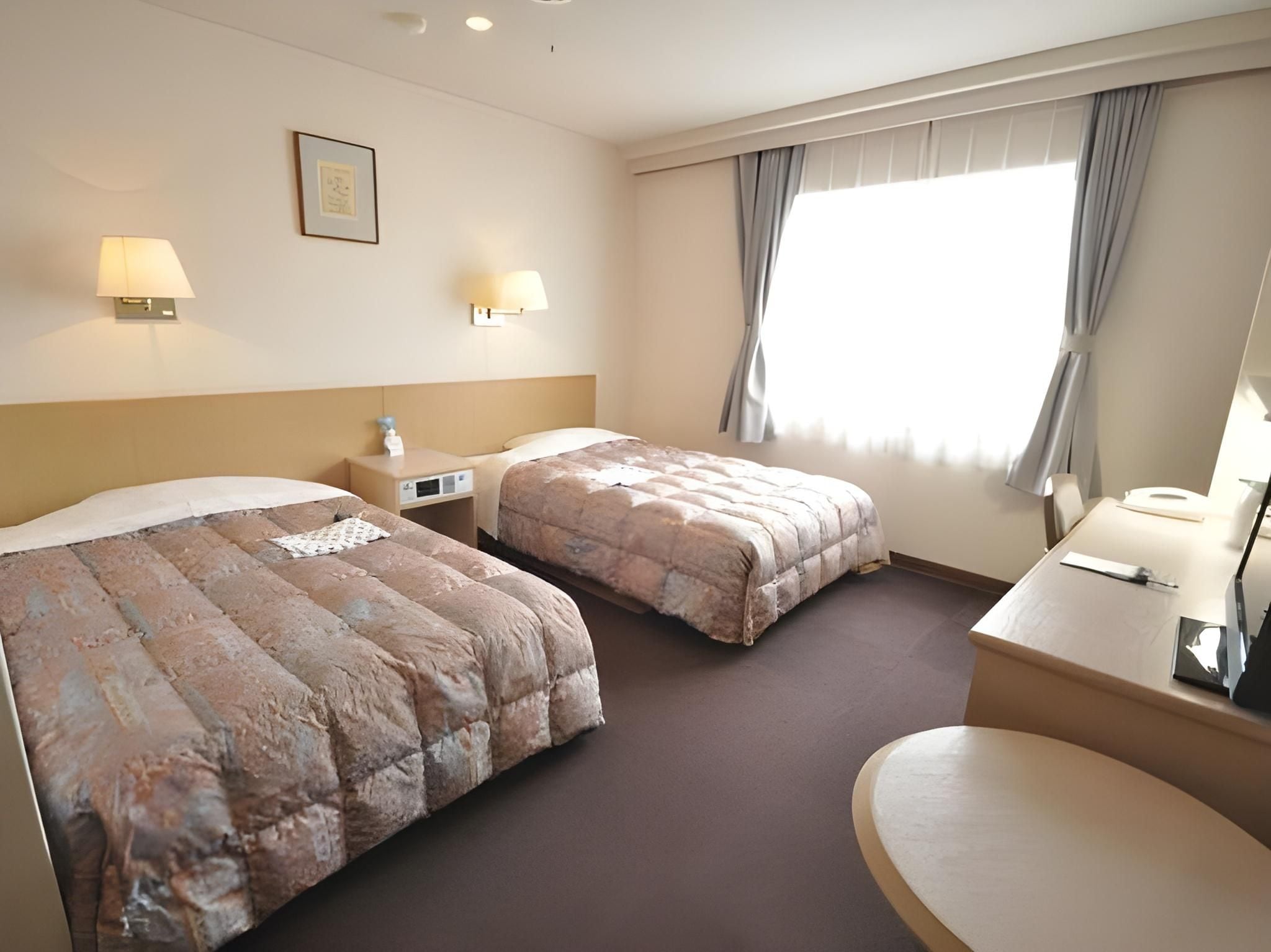 A hotel room with two beds, a desk, a chair, a lamp, and a window with curtains.