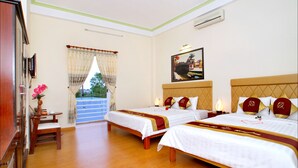 A hotel room with two beds, a desk, and a chair. There is a window with curtains, a painting on the wall, and a view of the outdoors. - Hoi An Life Homestay (Da Nang)