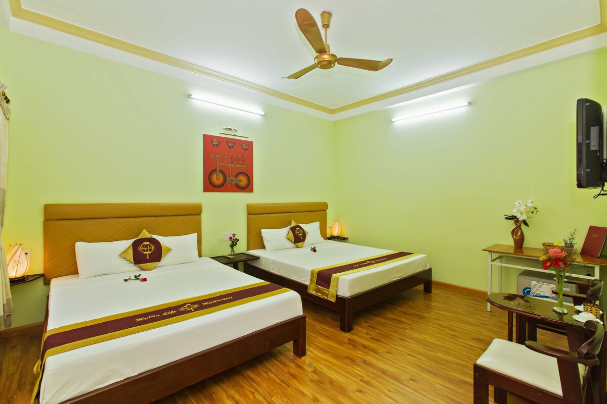 Une chambre d’hôtel avec deux lits, un ventilateur de plafond, un bureau avec une chaise et une petite table sur laquelle est posé un vase de fleurs.