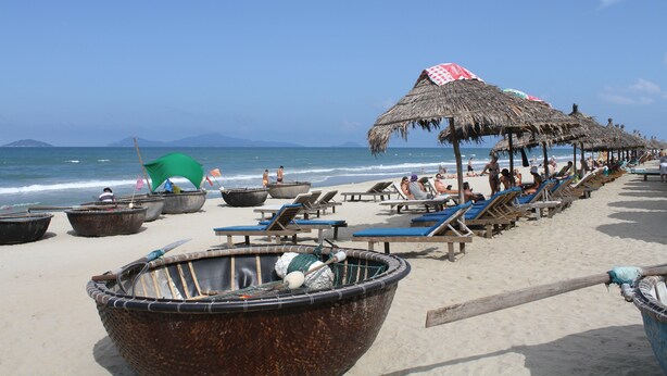A beach with thatched umbrellas, lounge chairs, and traditional boats. - Hoi An Life Homestay (Da Nang)