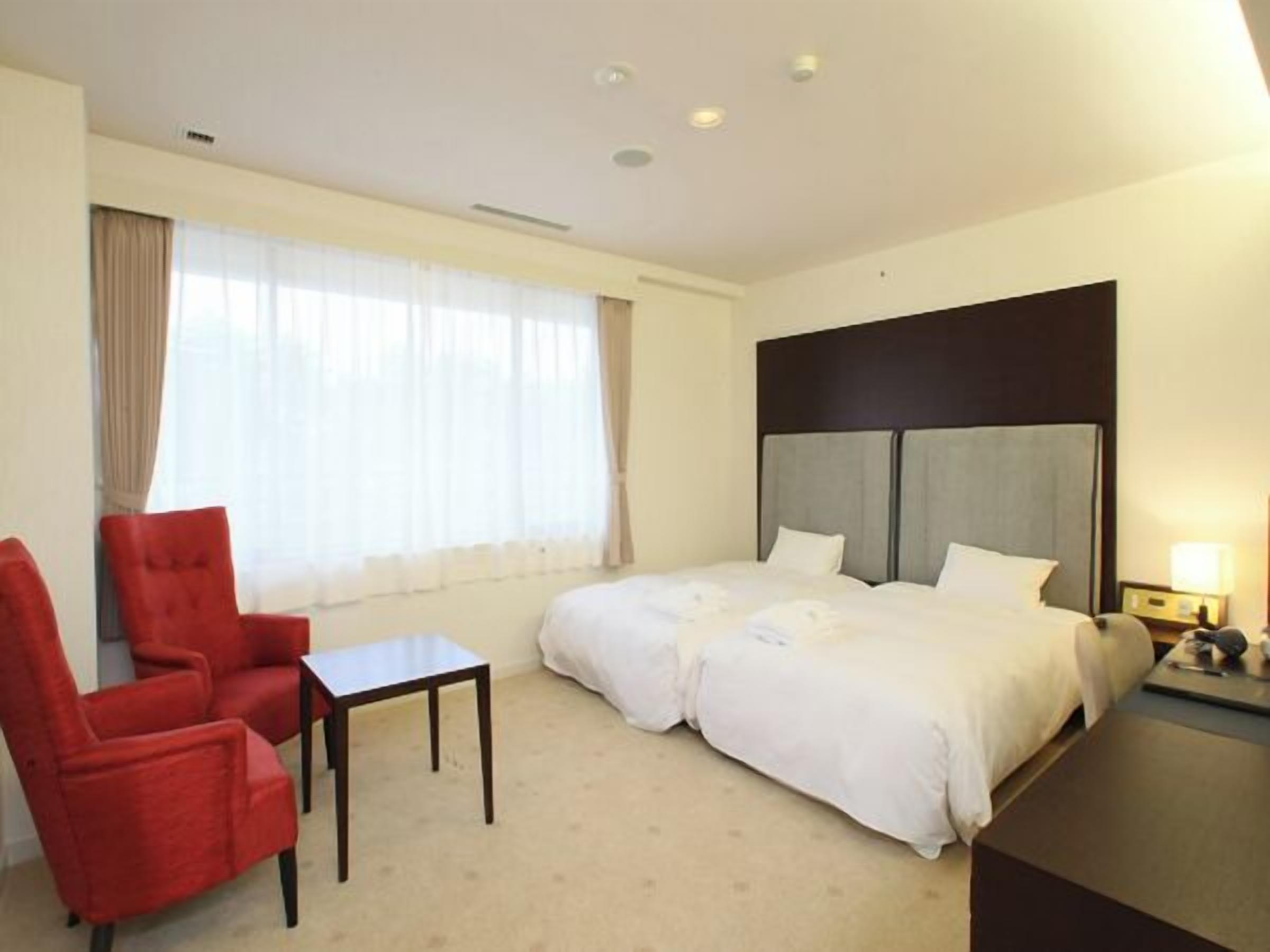 Ein Hotelzimmer mit Bett, rotem Sessel, kleinem Tisch und Nachttisch mit Lampe.