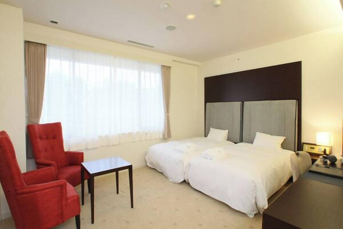 A hotel room with a bed, a red chair, a small table, and a nightstand with a lamp.