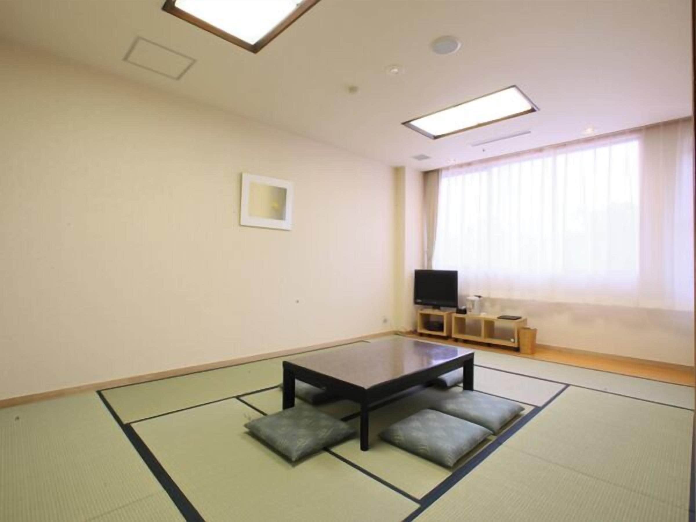 Ein Zimmer mit Tatami-Boden, einem niedrigen Tisch mit Kissen, einem Fernseher auf einem Schrank und einem Fenster mit Vorhängen.