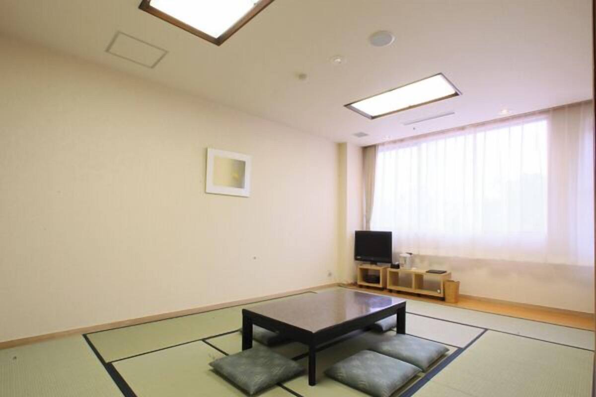 A room with tatami flooring, a low table with cushions, a television on a stand, and a window with curtains.