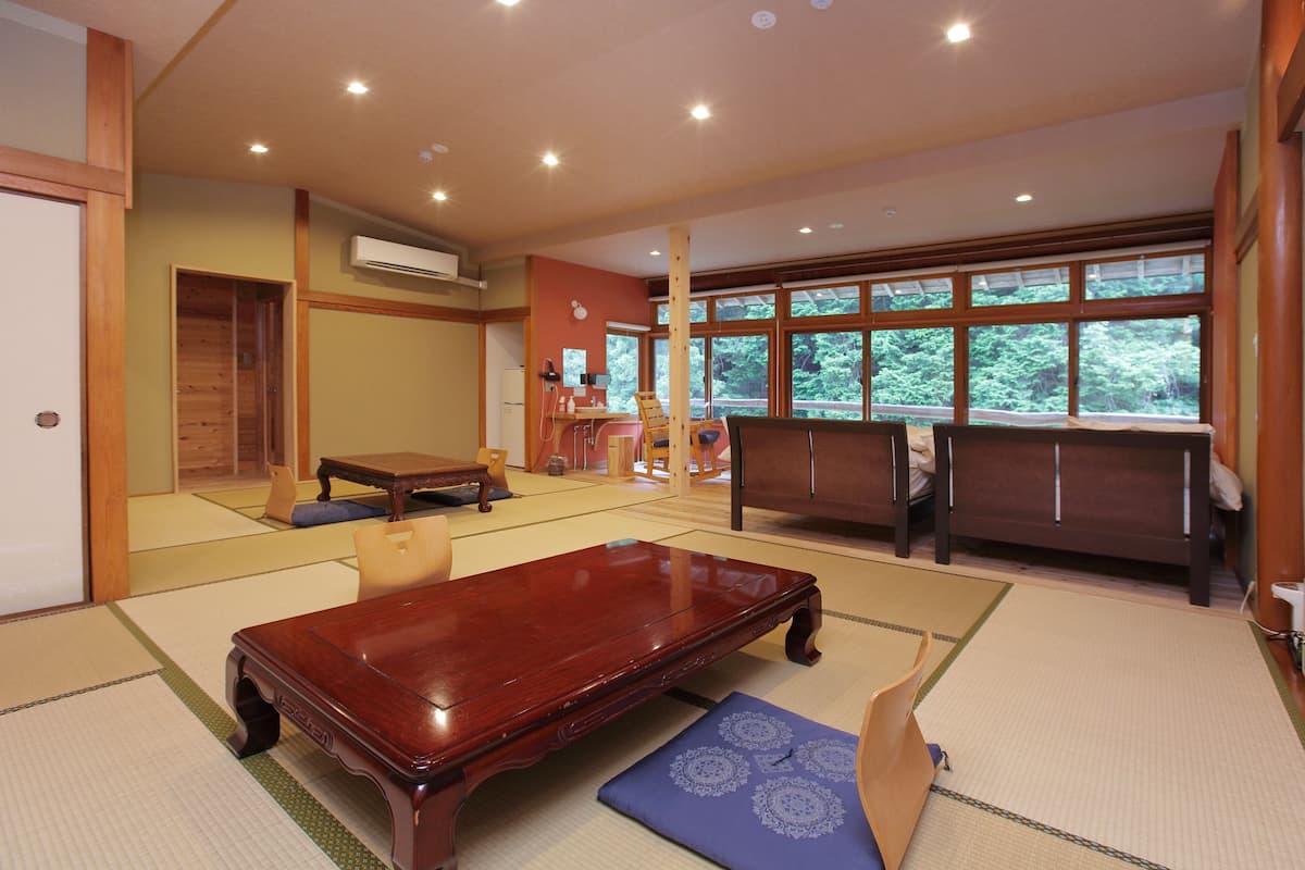 A traditional Japanese room with tatami flooring, a wooden table, and a view of greenery through large windows.