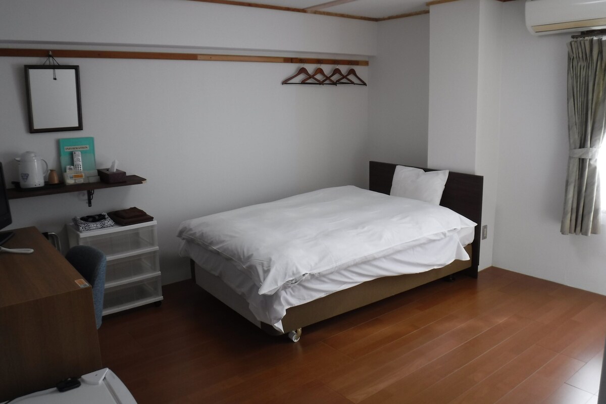 A bedroom with a bed, a desk, a chair, a shelf with items, and a window with curtains.