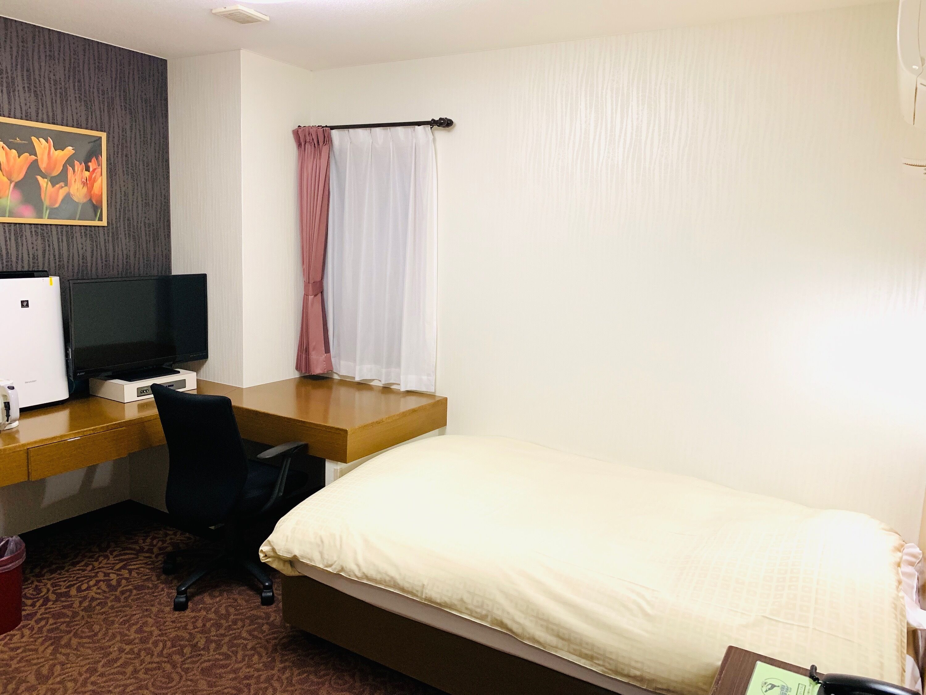 A hotel room with a bed, a desk with a computer, a TV, and a window with curtains.