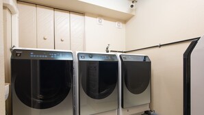 Une rangée de trois machines à laver à chargement frontal se trouve dans une buanderie, avec une porte blanche et un sèche-mains fixé au mur.