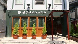 L’entrée d’un hôtel, avec une enseigne indiquant « 横浜タウンホテル » et « TOWN HOTEL ». Quatre plantes en pot sont disposées devant l’entrée.