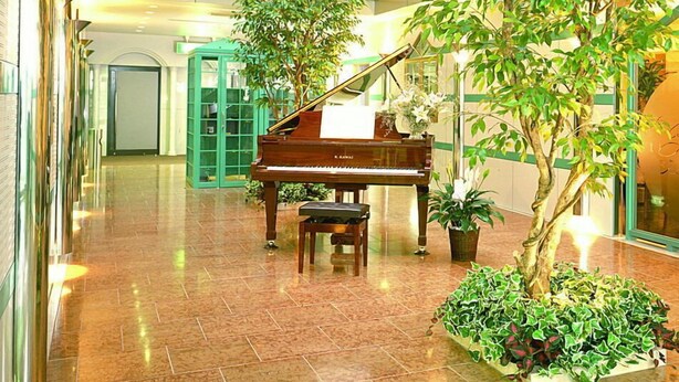 A grand piano in a spacious lobby with a high ceiling and glass panels, surrounded by potted plants and a tiled floor. - Ibaraki Central Hotel (Ibaraki)