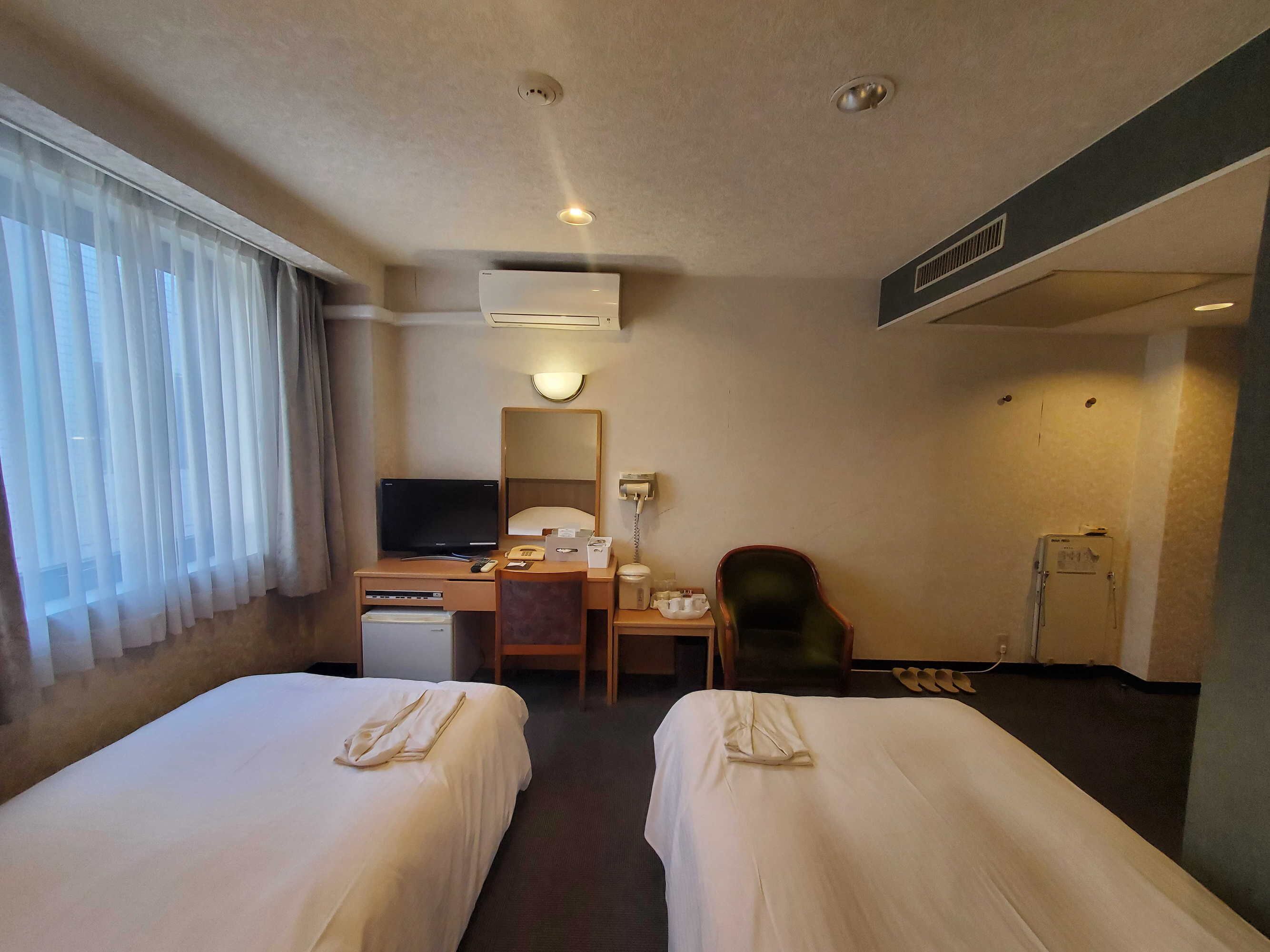 A hotel room with two beds, a desk with a computer, a chair, a TV, and a small table with a lamp.