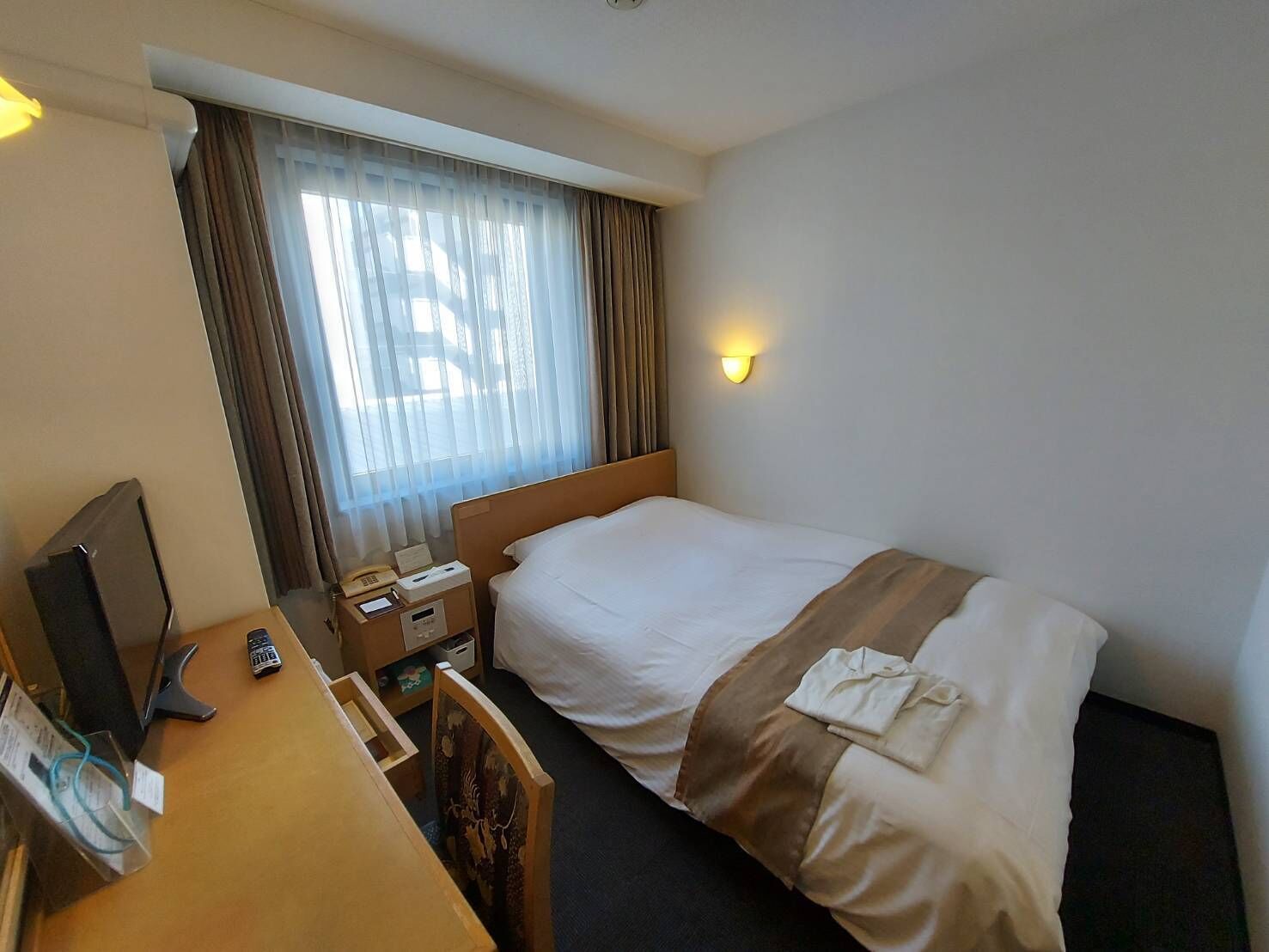 A hotel room with a bed, a desk, a chair, a nightstand, a lamp, and a window with curtains.