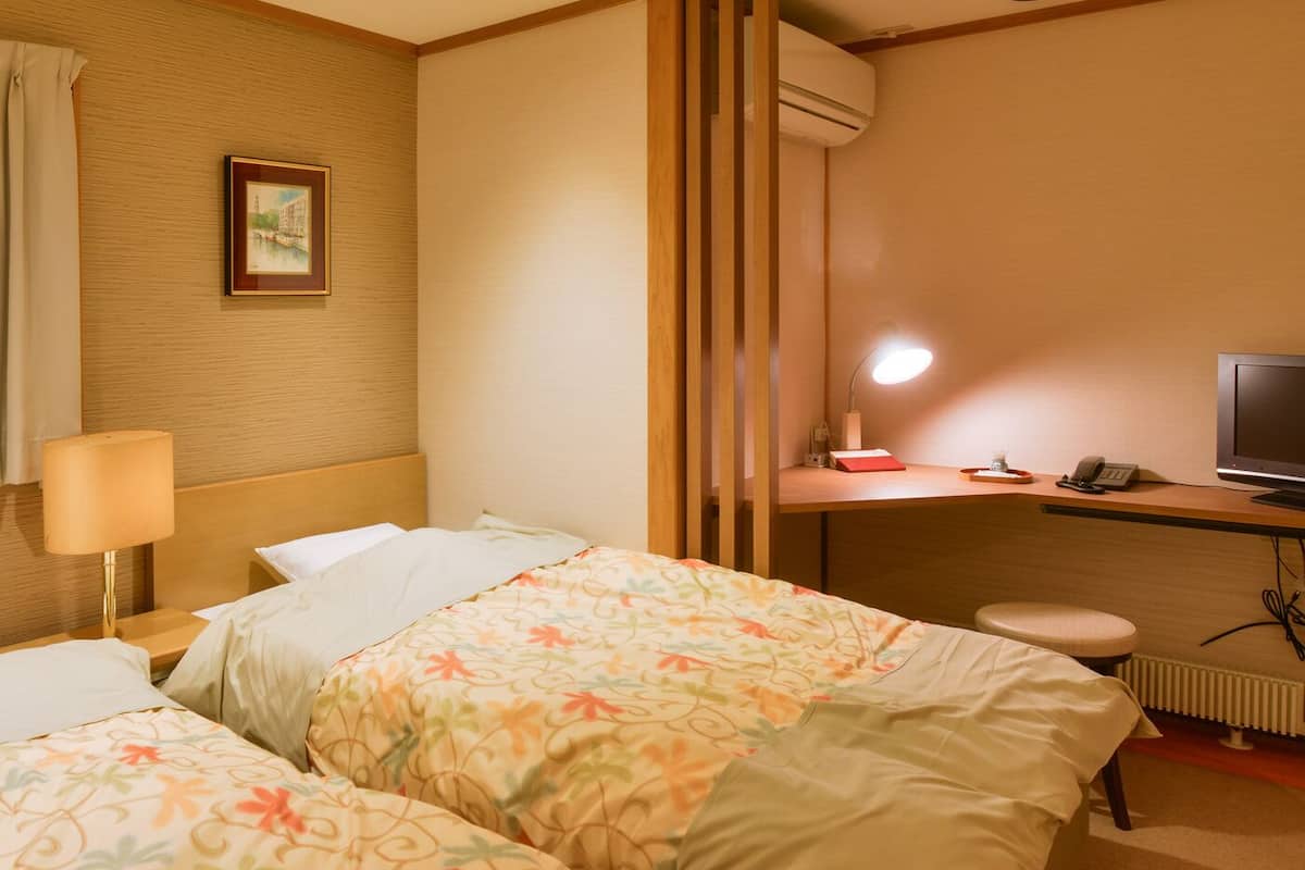 A hotel room with two beds, a desk with a lamp, a TV, and an air conditioning unit.