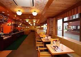 Interior de un restaurante con mesas y sillas de madera, una barra con la carta y vistas a la calle a través de amplias ventanas.